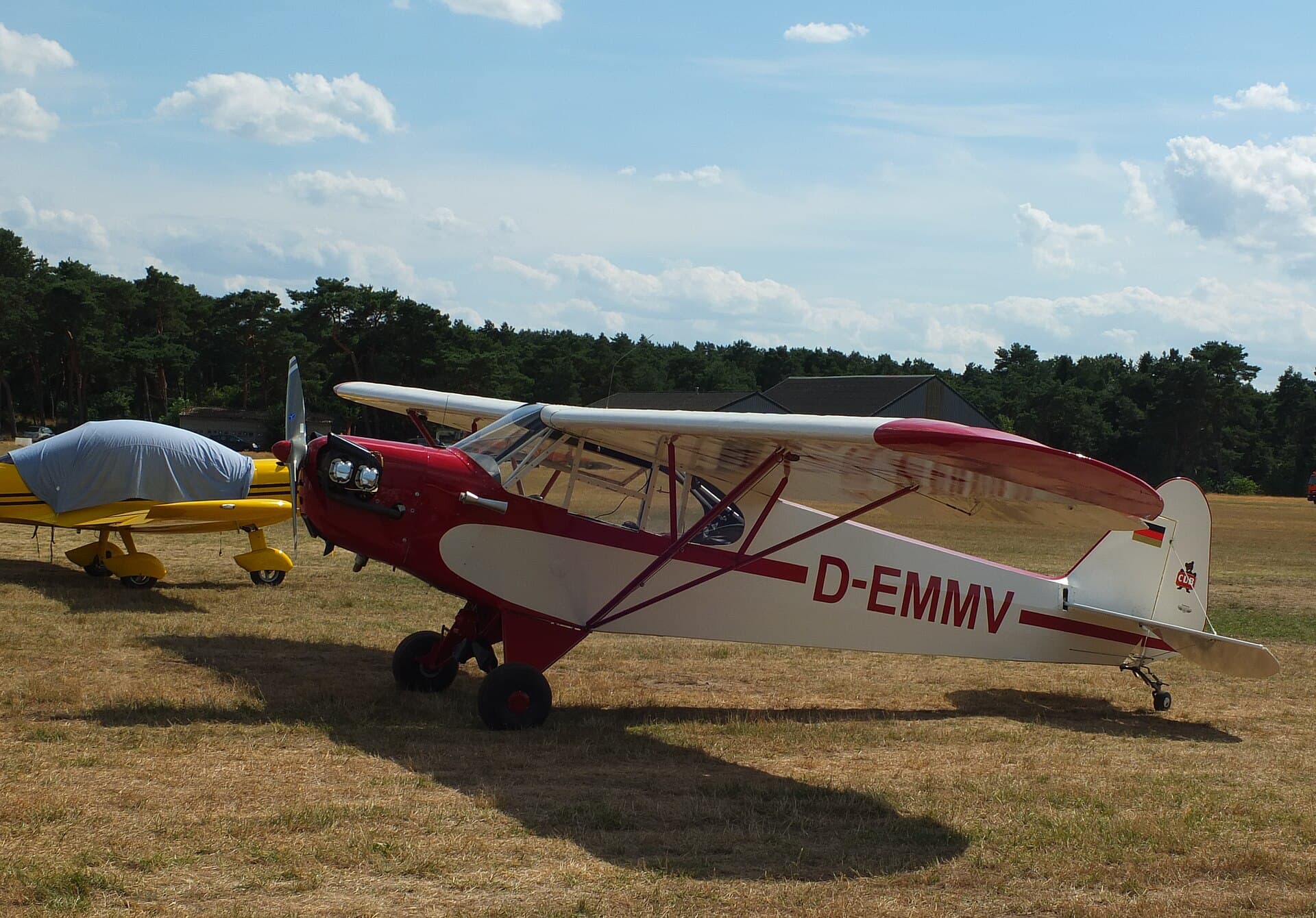 Un Piper Cub rouge et blanc, immatriculé D-EMMV, stationné sur un aérodrome herbeux