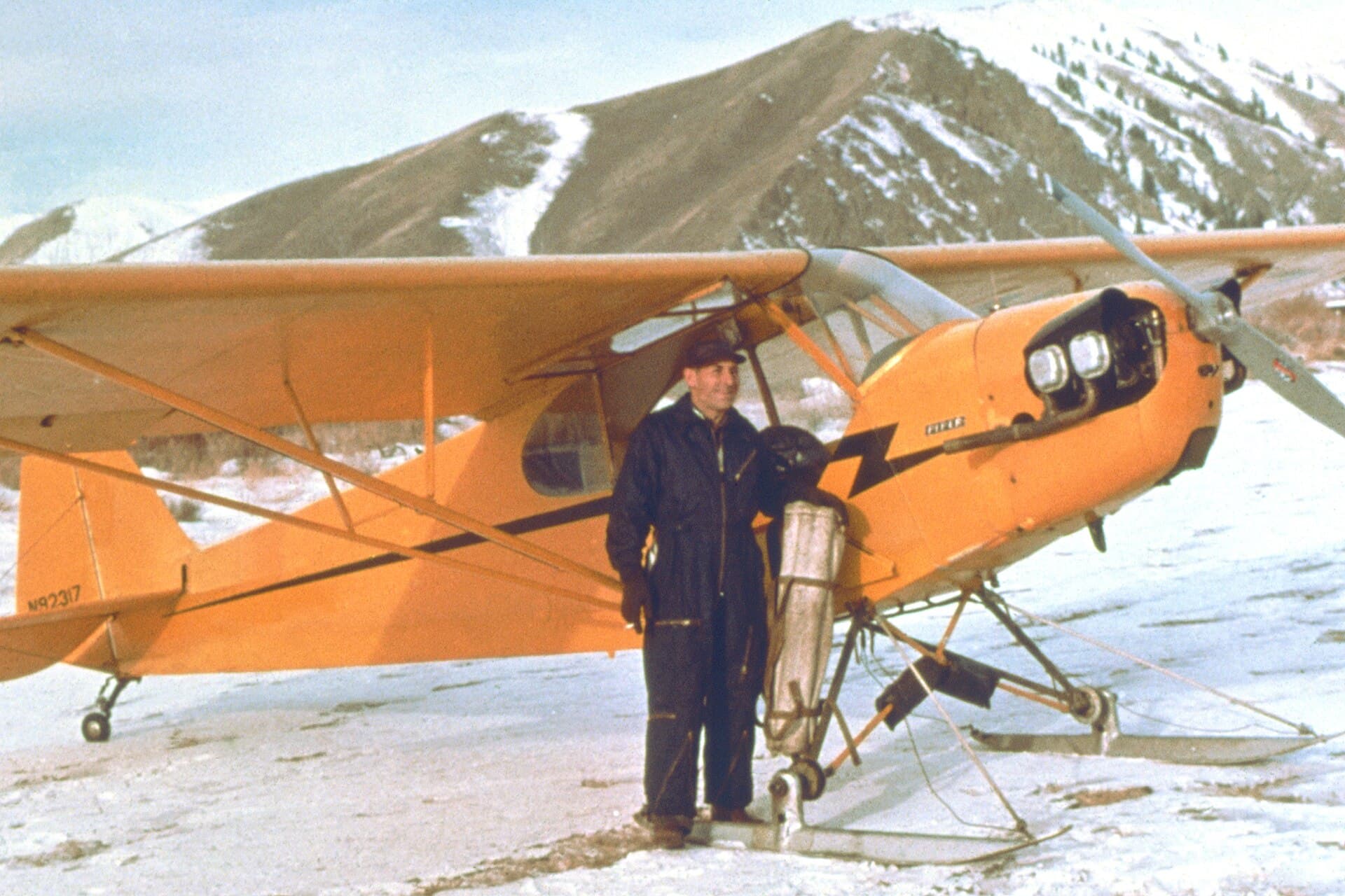 Un Piper Cub orange équipé de skis, posé sur la neige dans une vallée de montagne