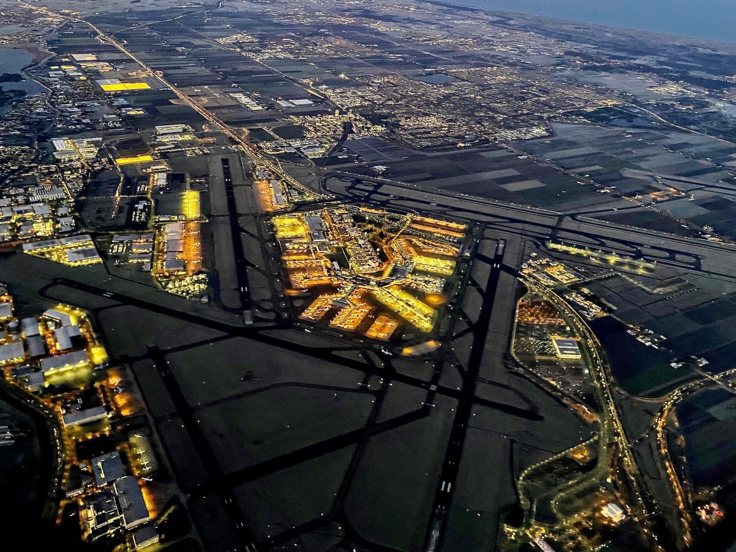 Schiphol à l'aube, vu d'un avion en montée : l'aérogare illuminée au centre de ses six pistes, étalées sur le polder