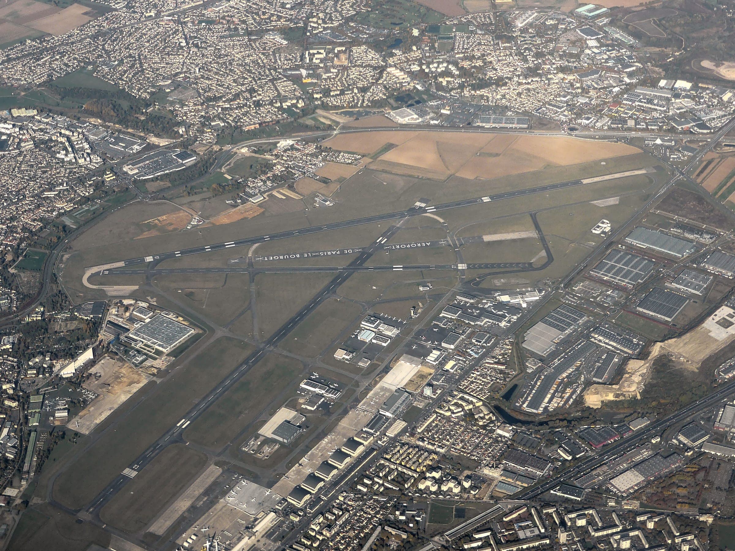 Vue aérienne plongeante du Bourget en automne, dans l'axe des pistes, avec les aires du musée, les hangars d'aviation d'affaires et l'habitat dense collé à la clôture