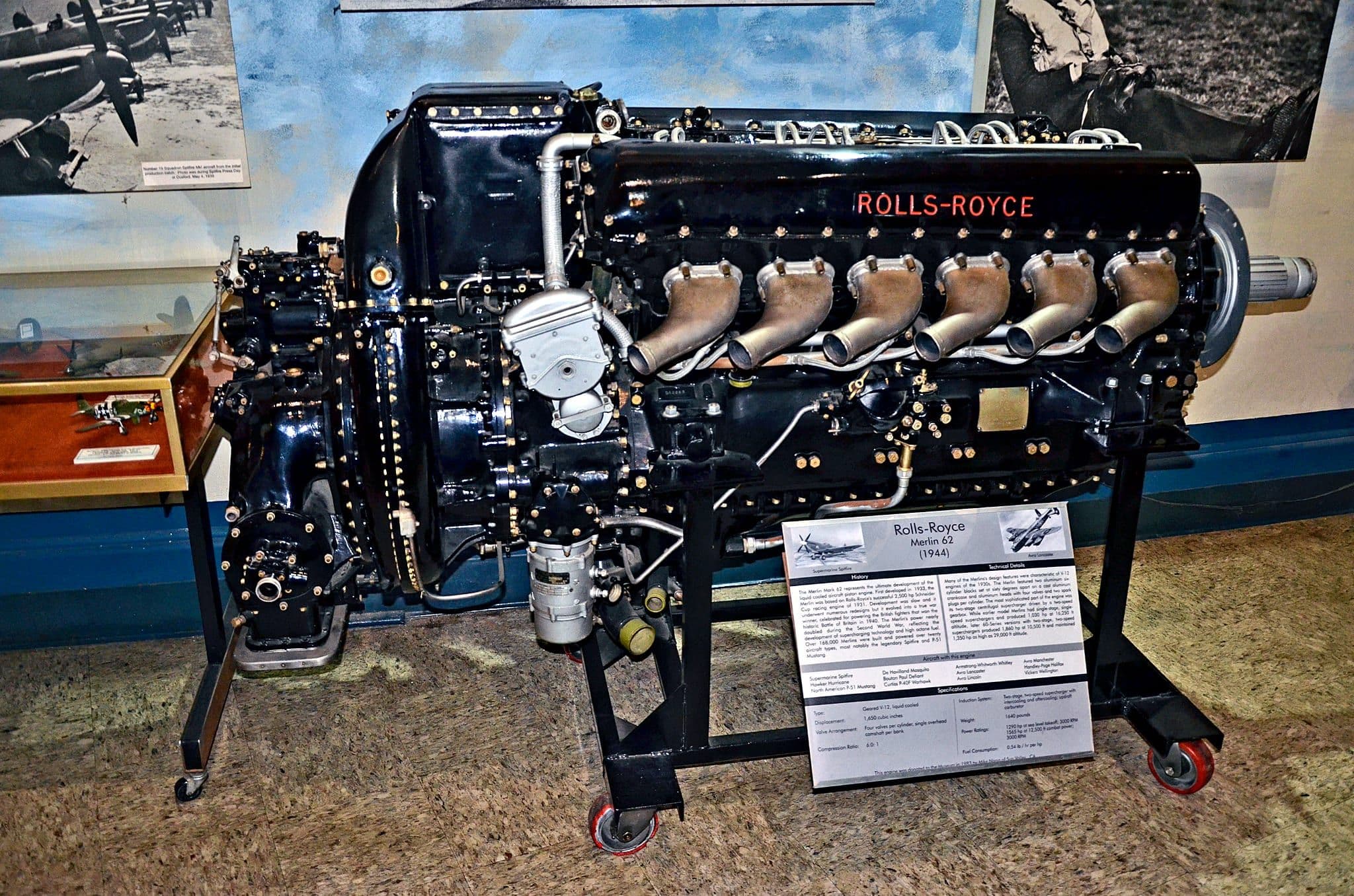 Un Rolls-Royce Merlin 62 de 1944 vu de profil sur un support de musée, blocs-cylindres noirs, tubulures d'échappement et carter de réducteur nettement éclairés