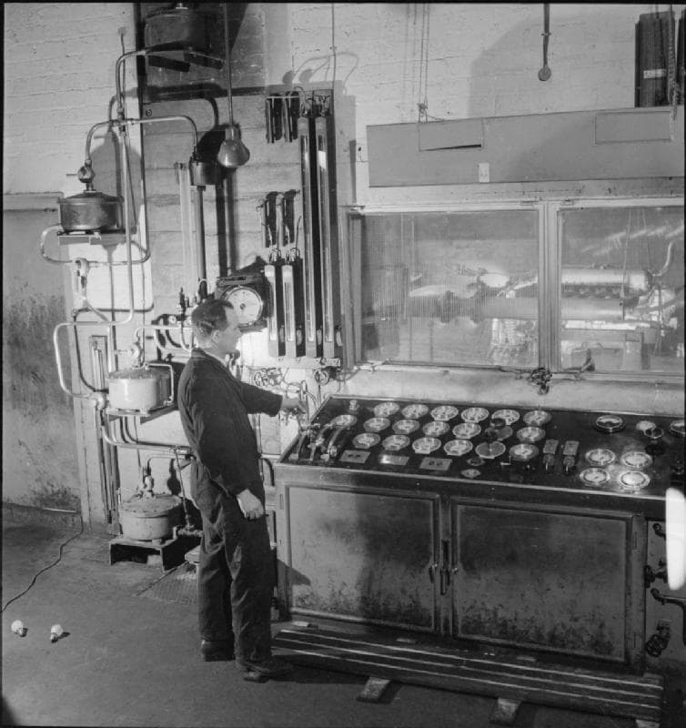 An engineer at the control panel of a Merlin test bed, the engine running behind the observation window, 1942