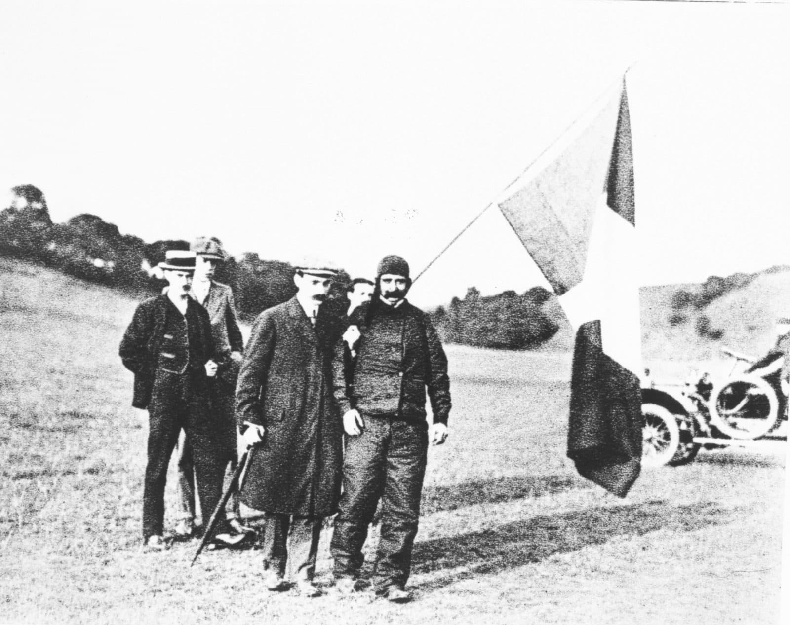 Trois hommes en manteau et casquette sur une pente herbeuse, près d'un drapeau et d'une automobile, catalogués comme Blériot après la traversée.