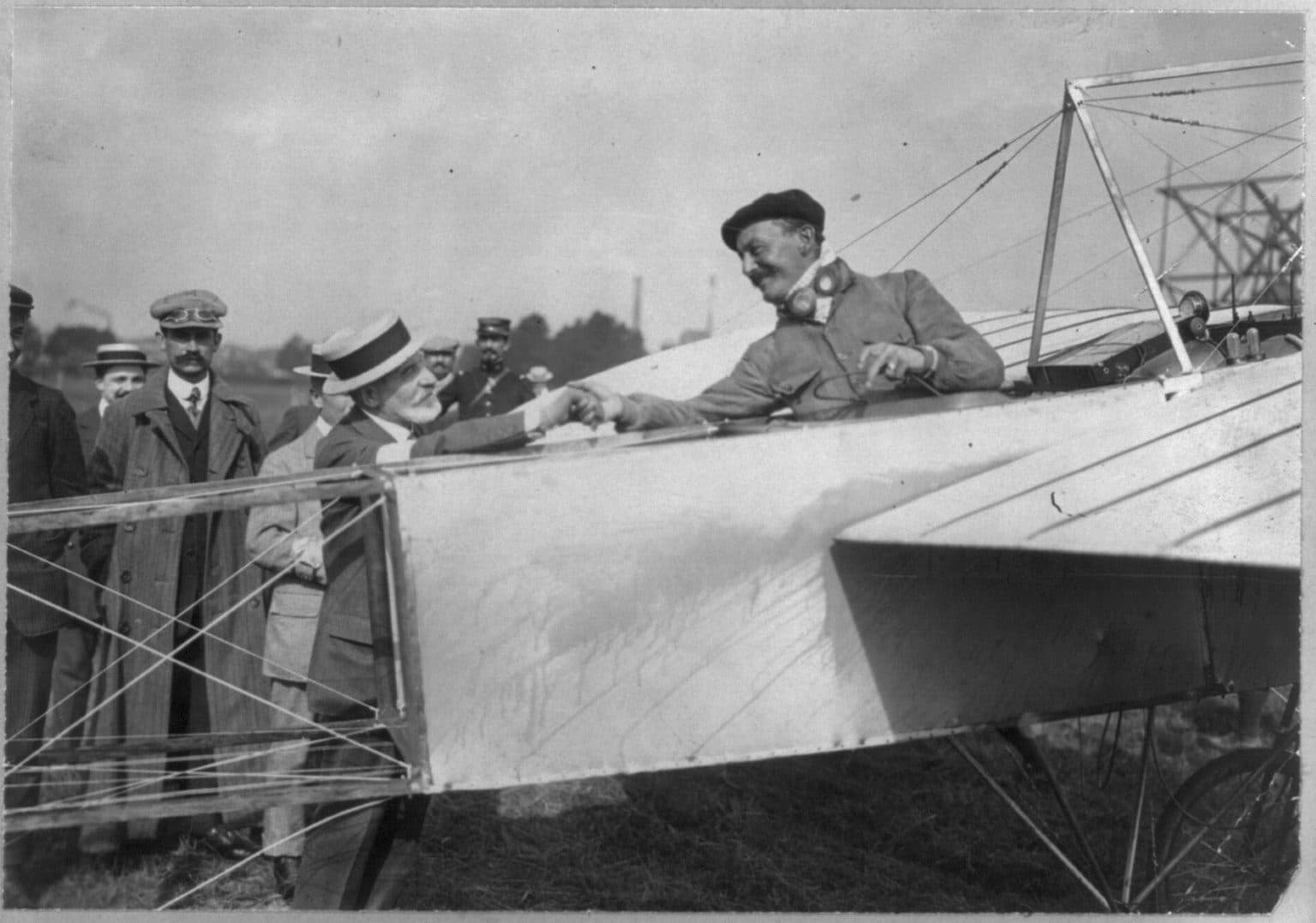 Alfred Leblanc, en casquette et manteau, penché sur le flanc d'un monoplan Blériot au milieu de la foule.