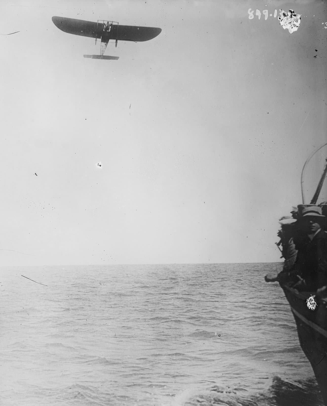 Un minuscule monoplan très haut au-dessus de la mer, le bastingage d'un navire en bord de cadre, catalogué comme Blériot au milieu de la Manche.