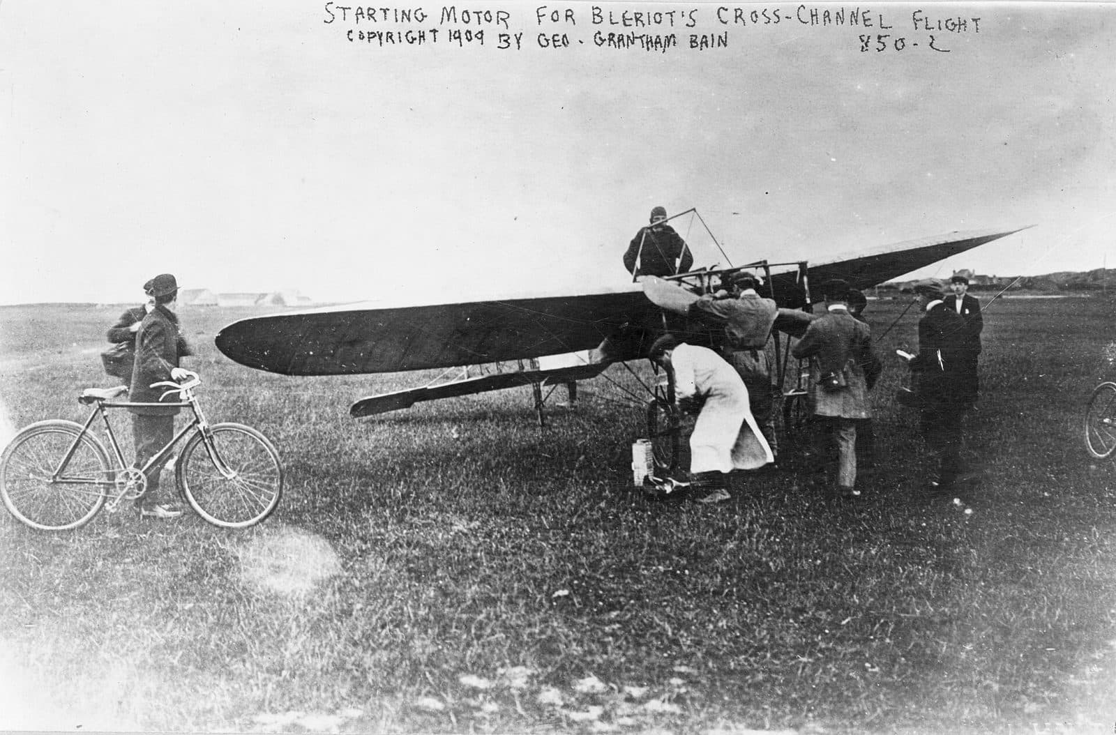 Le Blériot XI sur l'herbe des Baraques, des hommes penchés sur le moteur et un cycliste qui regarde, au moment où l'on lance le moteur pour la traversée.