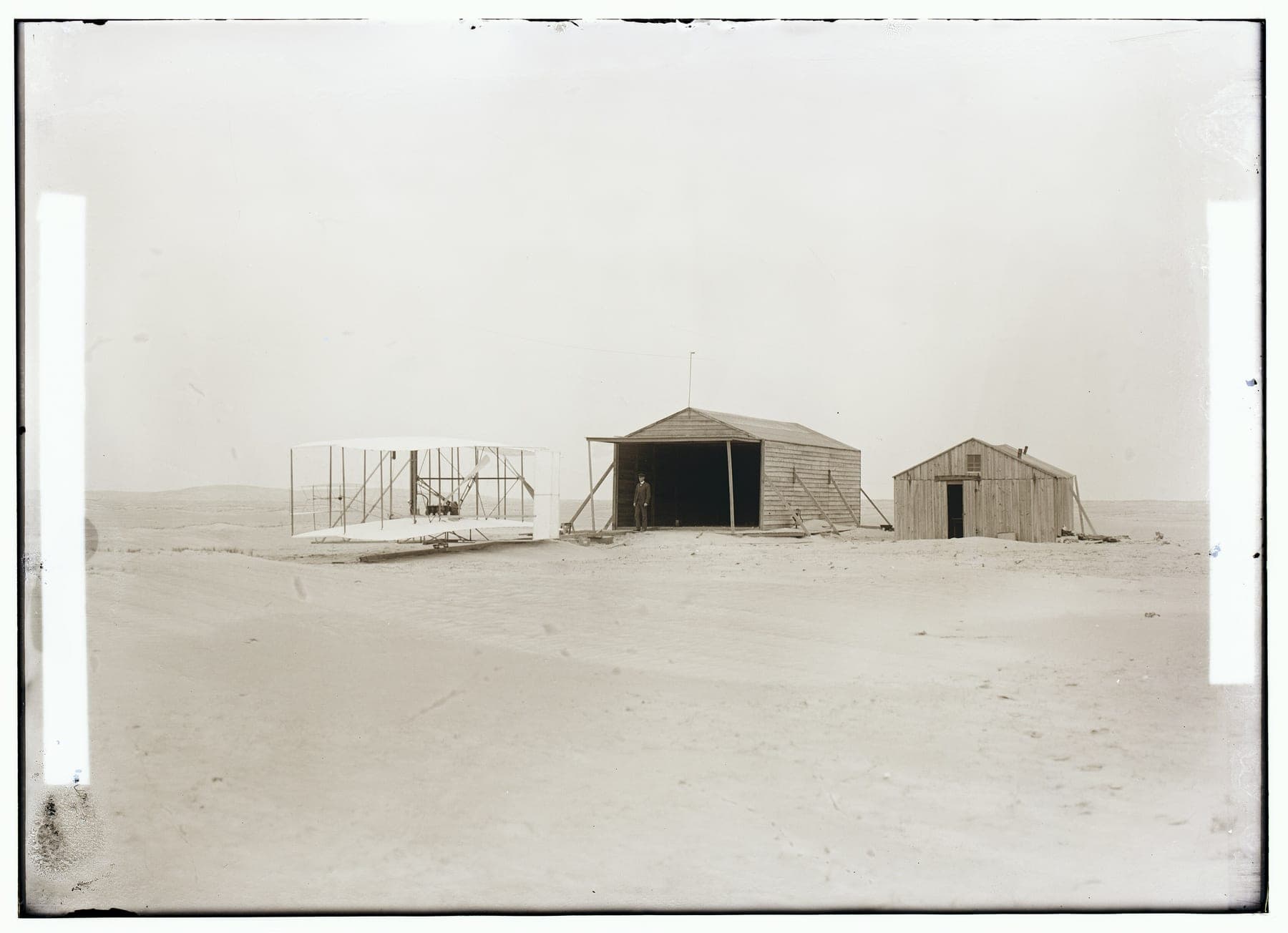 Le Flyer de 1903 devant les bâtiments du campement de Kill Devil Hills : un grand hangar abritant la machine et une cabane plus petite servant d'atelier et de logement.