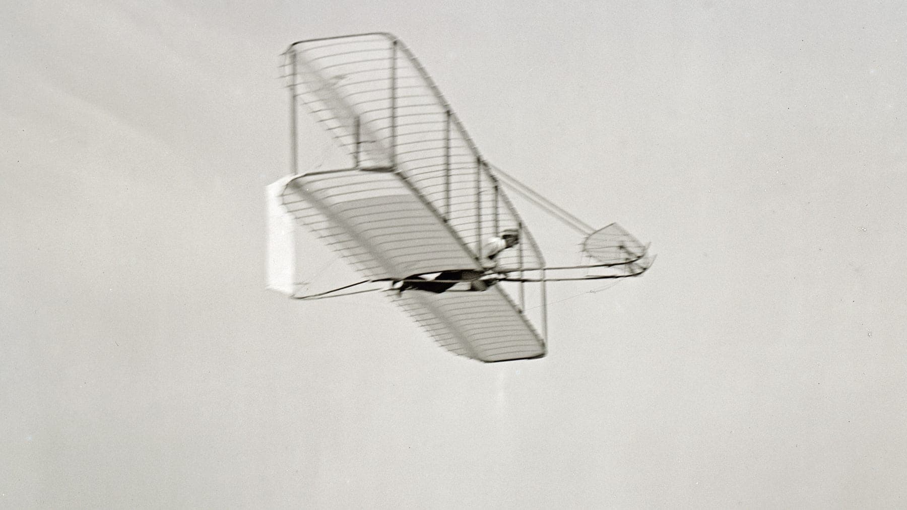Wilbur Wright en vol plané vers la droite sur la machine de 1902, vue de dessous, la dérive arrière mobile visible.