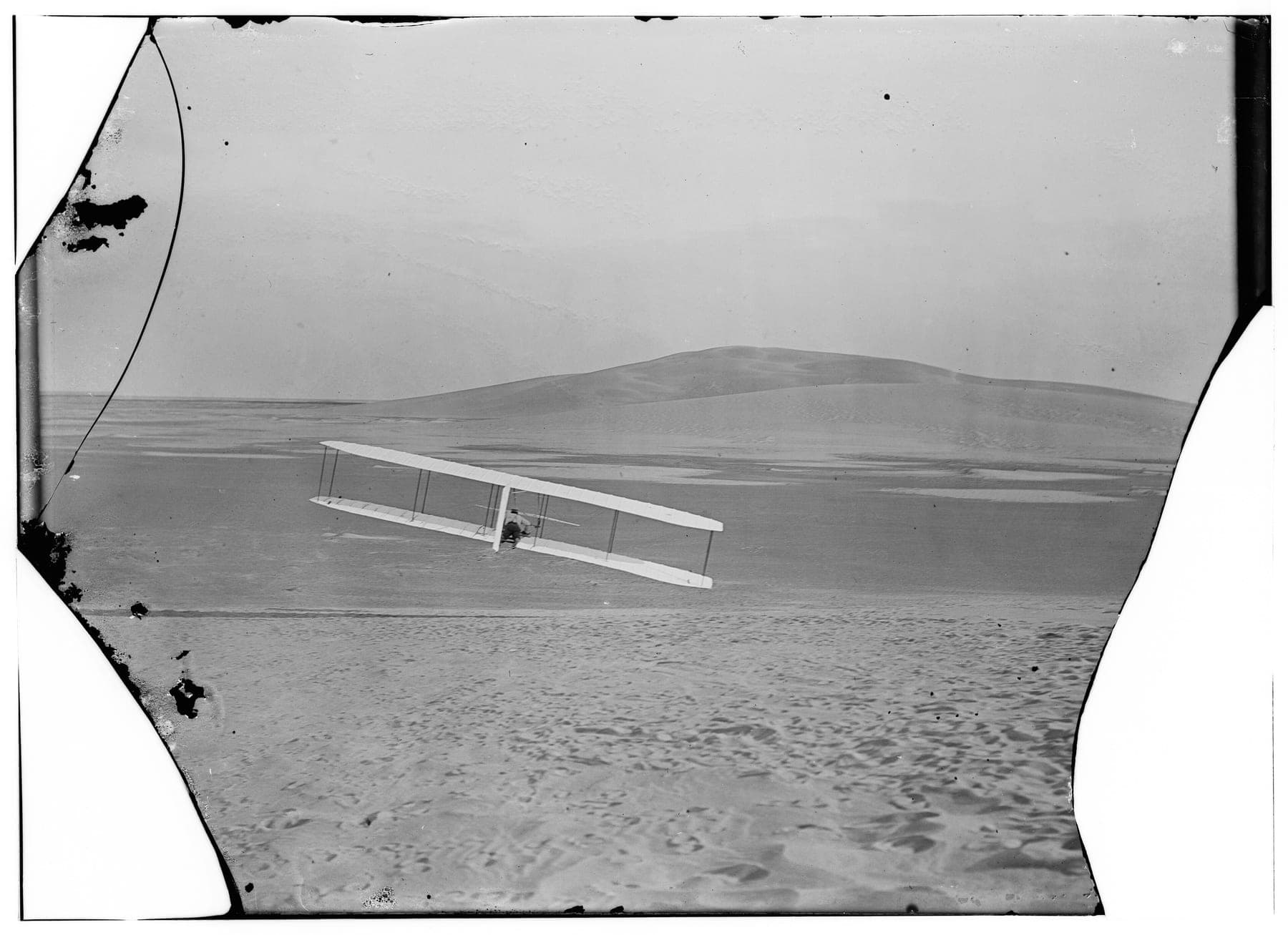 Le planeur de 1902 en virage à droite, vu de l'arrière, son extrémité d'aile droite près du sol.
