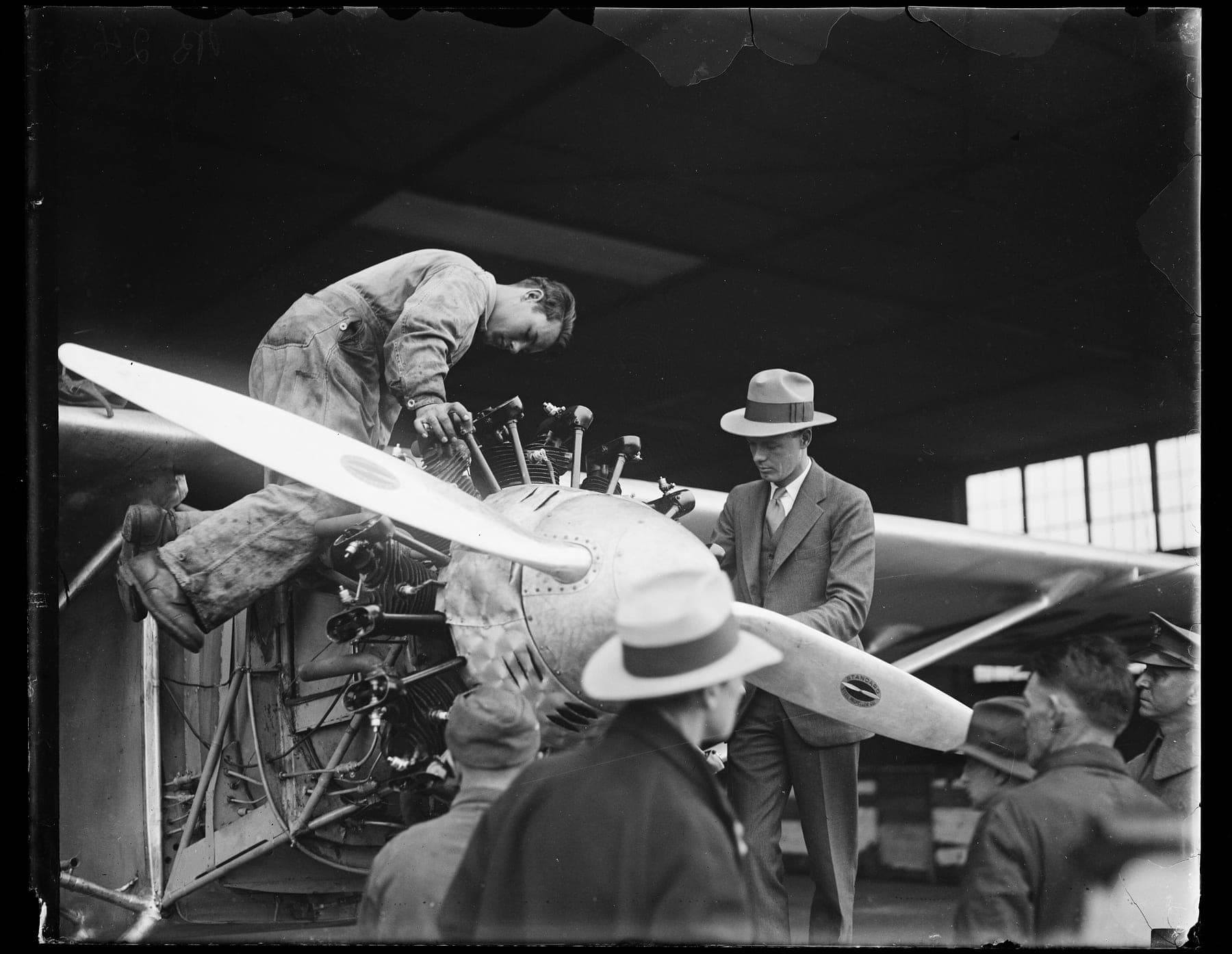 Lindbergh, en costume et chapeau, examine le moteur Whirlwind du Spirit pendant qu'un mécanicien travaille sur les cylindres.