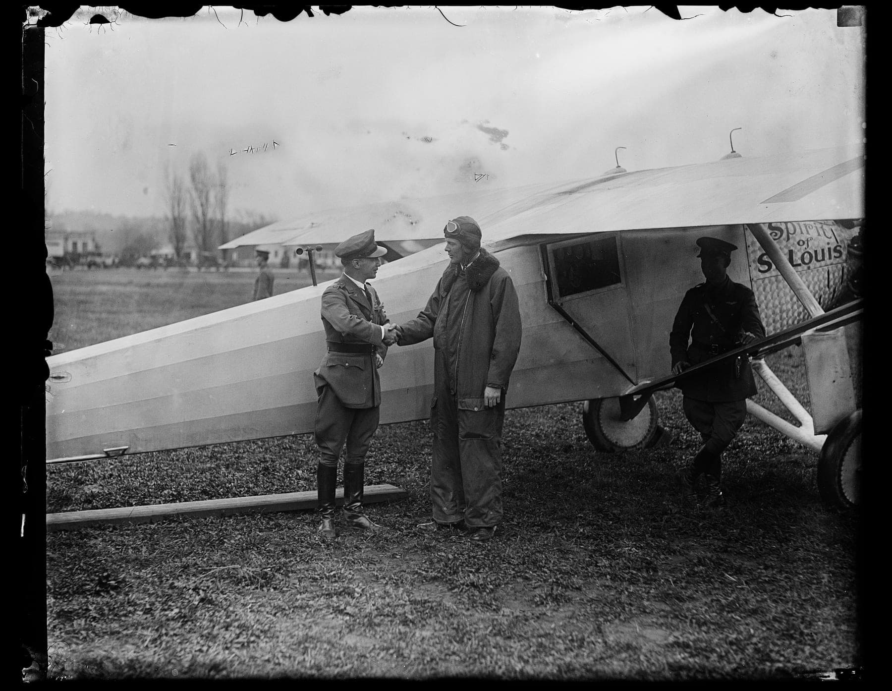 Charles Lindbergh, en combinaison et casque de vol, serre la main d'un officier de l'armée à côté du Spirit of St. Louis, dont le nom est peint sur le capot moteur.